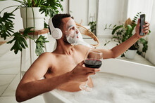Young Man With Facial Mask Drinking Wine And Making Selfie Portrait On Mobile Phone While Taking A Bath With Foam