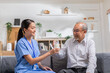 © 220 Selfmade studio - Asian beautiful therapist doctor serve milk to older patient in nursing home. Asian nurse in blue uniform assist elderly senior man to drink milk in hand while sitting at couch.Elderly care concept