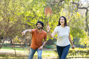 Happy cheerful young indian couple playing badminton game together in the park. urban asian sporty man and woman having fun outdoor sports and game activity concept. picnic and vacation.