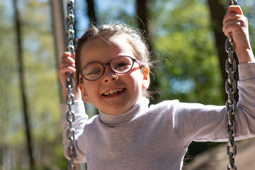 Little cute girl in glasses smiling rides on a swing at the playground in the park