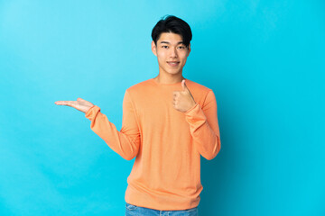 Wall Mural - Young Chinese man isolated on blue background holding copyspace imaginary on the palm to insert an ad and with thumbs up