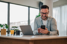 Distracted Smiling Businessman Using Smart Phone While Working At Office Desk