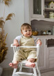 a baby boy in a beige jumpsuit is sitting in a white high chair in the kitchen. Child's diet