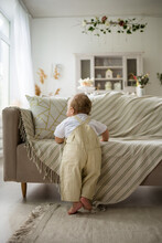 Caucasian Baby Boy In Beige Overalls Stands With His Back Near The Sofa In The Room
