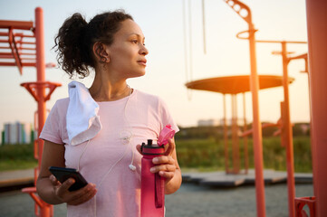 Athletic woman with smartphone and bottle of water resting after physical exercises on a sports ground with exercise equipment and crossbar at sunset