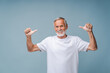 Happy senior man in white t-shirt shows thumbs-up gesture with hands smiling. Portrait of positive bearded grandfather standing on blue background