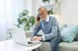 © Alla - Senior businessman discusses details of bargain with client on mobile phone. Elderly man works on new contract on laptop sitting at table at home