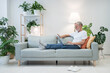 © Alla - Senior employee works online lying on soft couch against bright lamp and pot-plant at home. Focused elderly man types report in laptop leaning on cushions
