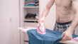 Masculine headless naked man in underwear home chore ironing blue shirt in bedroom