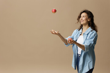 Wall Mural - Happy smiling focused curly beautiful female in jeans casual shirt juggling with apple posing isolated on over beige pastel background. Healthy food. Natural eco-friendly products concept. Copy space