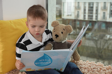 Sticker - Cute little boy with toy bear reading book near window at home