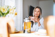 © Galina Zhigalova - Young beautiful happy woman with blonde long hair in white shirt drinking tea with bouquet of yellow flowers in vase on dinner table in bright interior at the home