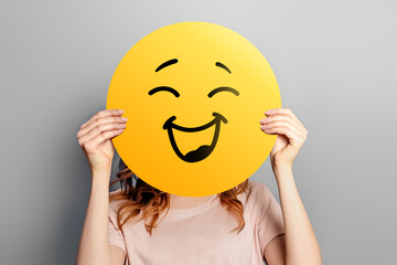 Wall Mural - Happy emoji. Girl holds a yellow emoticon with a smile isolated on a gray background. Happy emoji