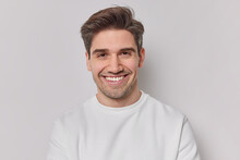 Portrait Handsome Man With Dark Hairstyle Bristle And Toothy Smile Dressed In White Sweatshirt Feels Very Glad Poses Indoor. Pleased European Guy Being In Good Mood Smiles Positively. Emotions Concept