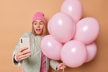 Wall Mural - Beautiful young woman looks surprisingly at front camera of smartphone takes selfie wears pink hat and jacket holds bunch of pink inflated balloons enjoys party time isolated over brown background