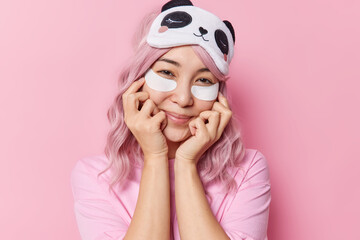 Wall Mural - Portrait of charming Asian woman keeps hands under chin smiles pleasantly applies white beauty pads under eyes wears casual t shirt and blindfold isolated over pink background. Skin treatment