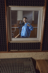 Wall Mural - Smiling African young woman singer shows thumb up standing near microphone inside a soundproof room of recording studio