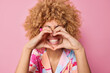 Positive curly haired young woman makes heart gesture over toothy smile has good romantic mood dressed casually expresses love to someone isolated on pink background. My heart belongs to you