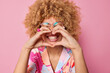 Happy curly haired young woman makes heart gesture over mouth smiles toothily wears transparent spectacles and colorful shirt says I love you isolated over pink background. Body language concept