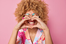 Happy Curly Haired Young Woman Makes Heart Gesture Over Mouth Smiles Toothily Wears Transparent Spectacles And Colorful Shirt Says I Love You Isolated Over Pink Background. Body Language Concept