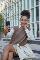 Wall Mural - Vertical shot of happy curly haired teenage girl shoots video content for sharing in social media networks enjoys travel vacation dressed in casual clothes poses outdoors on bech at urban area