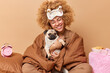 © WHstudio Leushin N - Happy woman wrapped in blanket holds pug dog expresses love to favorite pet has glad expression poses on comfortable bed alarmclock bowl of cornflakes around beige background. Time to having rest