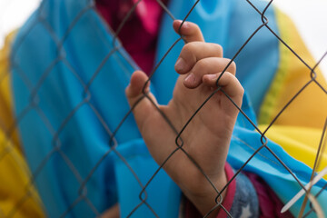 A little refugee girl with a sad look behind a metal fence. Social problem of refugees and internally displaced persons. Russia's war against the Ukrainian people