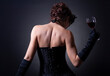 © papa - A young woman in a corset and long gloves with a glass of red wine stands with her back to the viewer.