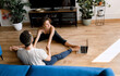 © shunevich - Sporty young couple warming up stretching watching fitness video tutorial online on laptop, fit man and woman doing workout at home sitting on floor in living room practicing yoga and holding hands