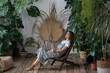 © DimaBerlin - Young calm hipster woman relaxing sit on comfortable armchair in stylish scandinavian indoor garden with green exotic plants. Happy florist girl enjoy spending time with houseplant in home greenhouse