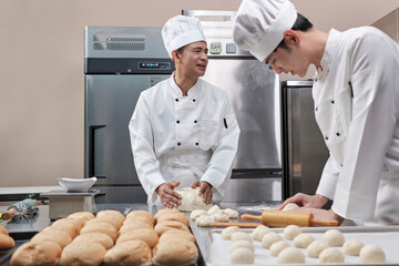 Two professional Asian male chefs in white cook uniforms and aprons are kneading pastry dough and eggs, preparing bread and fresh bakery food, baking in oven at stainless steel kitchen of restaurant.