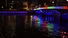 Colorful Bridge Reflects Off Ripples In River Through City At Night