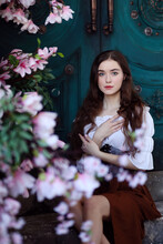 Beautiful Long Haired Girl In Medieval Dress Sitting On Steps In Front Of Blue Door Surrounded By Magnolia Flowers