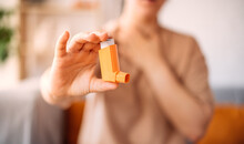 A Girl Suffering From An Asthma Attack Due To Allergies Shows An Inhaler While Sitting On The Sofa In The Living Room Of The House. The Concept Of Combating Allergies. Selective Focus
