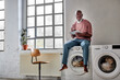 © Westend61 - Bald man using tablet PC sitting on washing machine at home