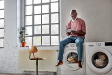 Bald Man Using Tablet PC Sitting On Washing Machine At Home