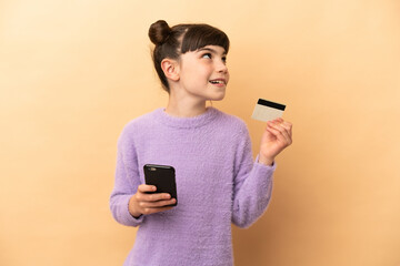 Wall Mural - Little caucasian girl isolated on beige background buying with the mobile with a credit card while thinking