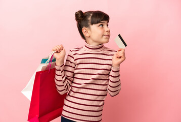 Wall Mural - Little caucasian girl isolated on pink background holding shopping bags and a credit card and thinking