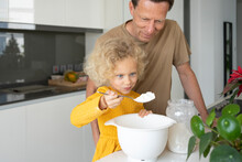 Smiling Blond Girl Holding Spoon Of Flour Standing By Father In Kitchen