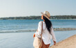 © Syda Productions - people, summer holidays and leisure concept - happy young woman in white shirt and straw hat with bag walking along beach