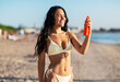 © Syda Productions - people, summer and swimwear concept - happy smiling young woman in bikini swimsuit using sunscreen spray on beach