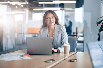 Smiling business lady wearing eyeglasses working on laptop in the corporate company at modern office