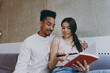 © ViDi Studio - Two young friends woman man of African American ethnicity in casual clothes read book write down in diary sitting on gray sofa indoor spend free time rest in flat together. People lifestyle concept.