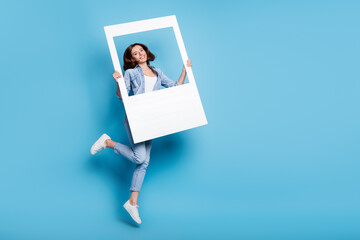 Poster - Photo of shiny cute woman wear jeans shirt holding white frame jumping empty space isolated blue color background