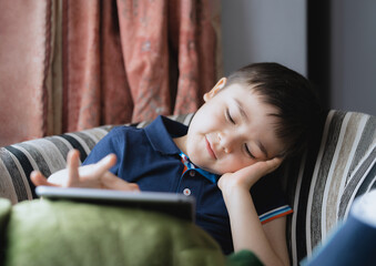 Wall Mural - Cinematic portrait young boy playing game on tablet sitting on sofa with light shining from window, Kid playing games online on internet at home, Child talking video call with friends at home