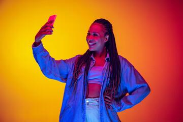 Wall Mural - Young pretty girl in cotton shirt using phone isolated on yellow red background in neon light. Concept of beauty, art, fashion, youth and emotions