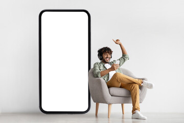 Joyful indian guy sitting in armchair with smartphone and headset