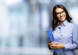 © vgstudio - Happy smiling businesswoman with blue folder with documents. Portrait image - young brunette business woman in eye glasses spectacles, indoor, over blurred modern office interior background.