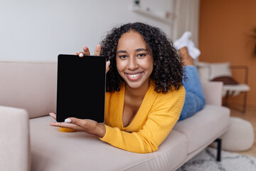 Poster - Happy young African American woman showing tablet computer with blank screen, lying on sofa at home, mockup
