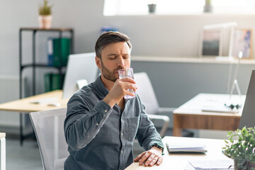 Poster - Mature male manager holding glass of water, drinking water while working on computer in office, copy space